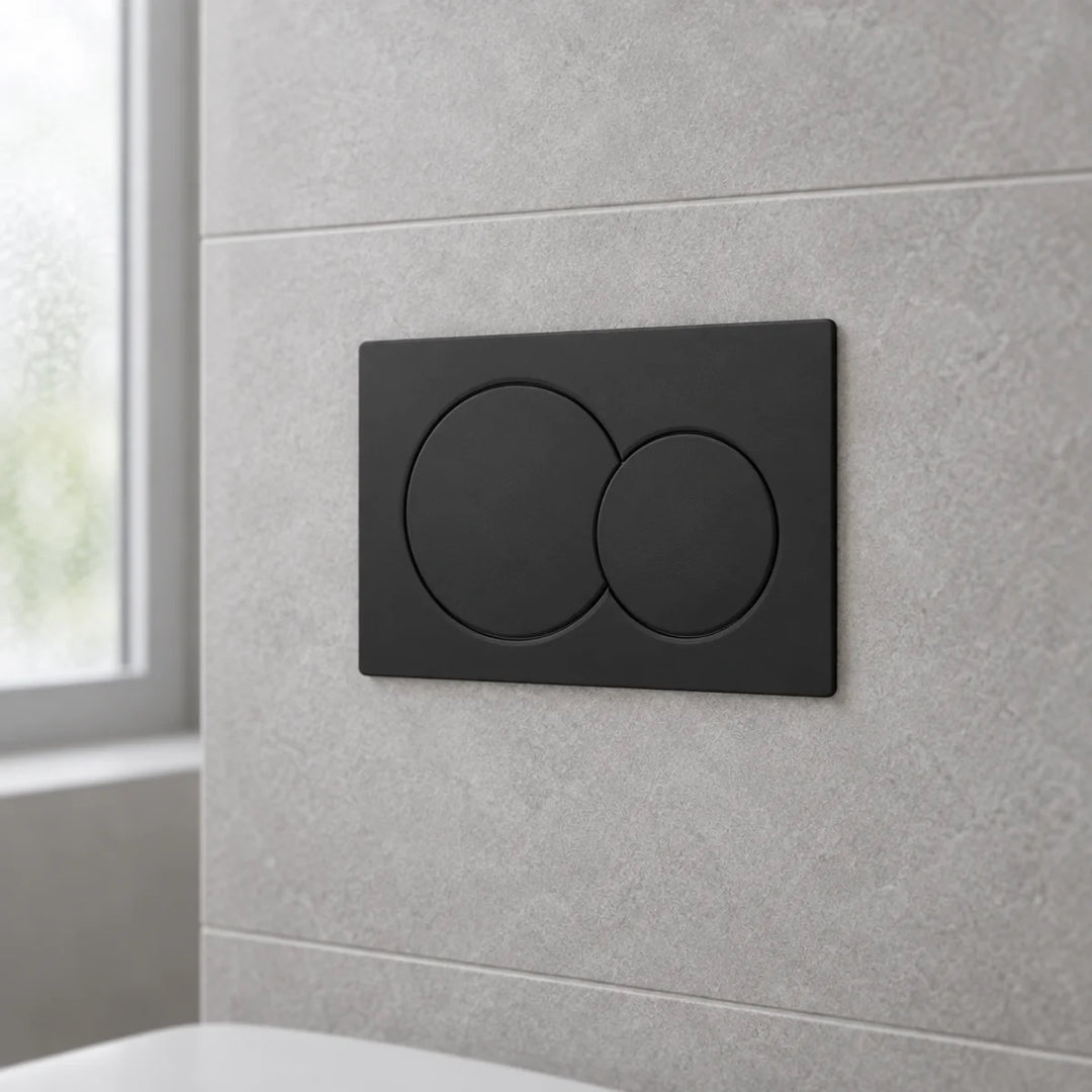 Geberit Alpha01 round dual flush plate in matt black, angled view mounted on a grey tiled bathroom wall beside a window