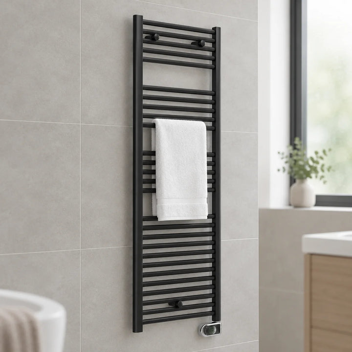 Essential Electric ERP black heated towel rail with a white towel, wall-mounted on grey tiles in a bathroom with window and vanity