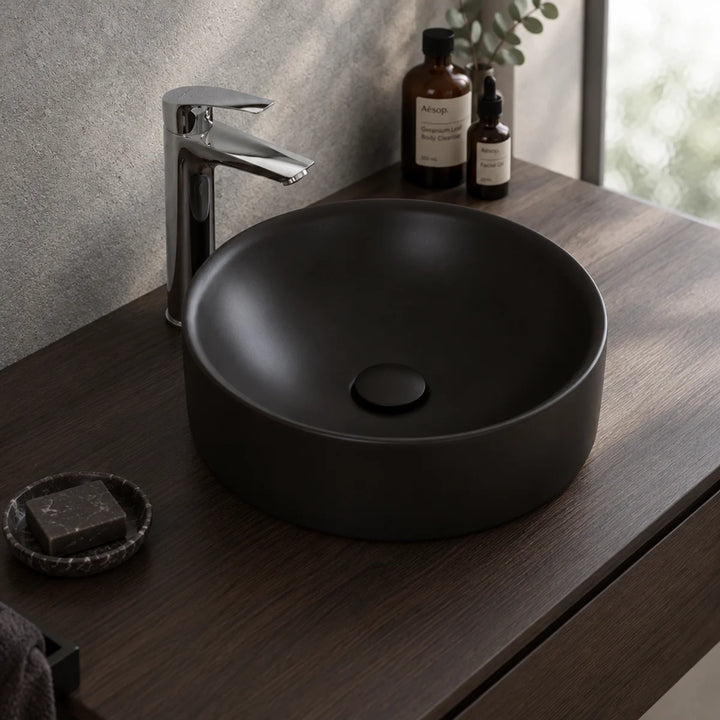 Duravit Durapoint round matt black countertop washbowl on a dark wood vanity with a chrome tall mixer tap, soap dish and toiletries beside it