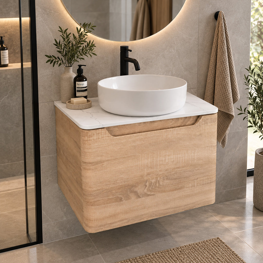 A modern wall-mounted bathroom vanity with a light oak wood cabinet, white marble countertop, and a white oval vessel sink. The vanity is styled in a contemporary bathroom with a gray tiled wall, a black faucet, a round backlit mirror, a beige towel, and small decorative plants.