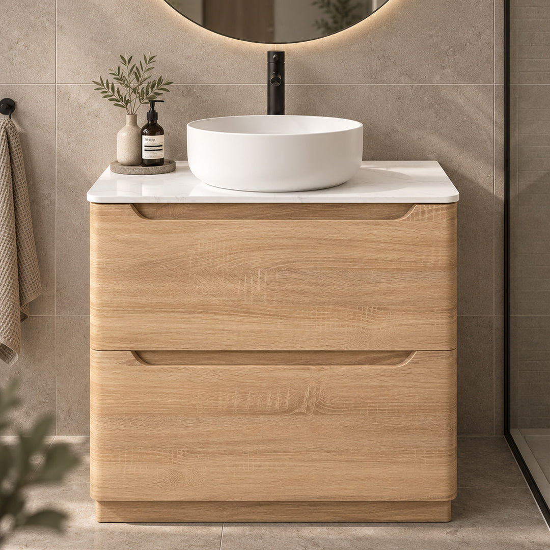 A modern bathroom vanity with a light oak wood cabinet, a white marble countertop, and a white oval vessel sink. The vanity is styled in a contemporary bathroom with a beige tiled wall, a black faucet, and a round backlit mirror.