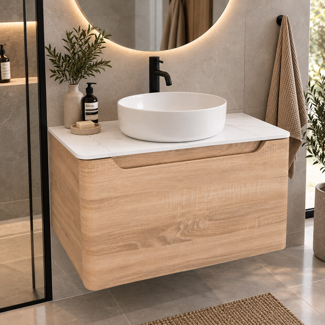 A modern wall-mounted bathroom vanity with a light wood cabinet, white marble-look countertop, and a white oval vessel sink. The vanity is shown in a styled bathroom setting with a black faucet, a lit round mirror, a towel, and small decorative plants.