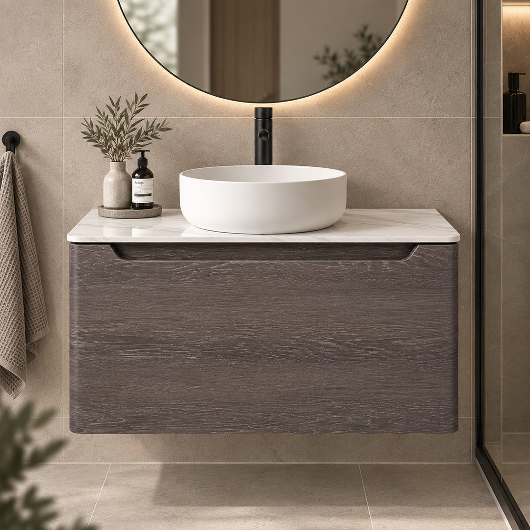 A modern floating bathroom vanity with a dark wood grain cabinet, a white marble countertop, and a white oval vessel sink. The vanity is styled in a bathroom setting with a round backlit mirror, a black faucet, and decorative items on the counter.