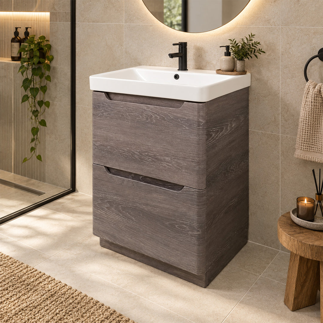 A modern bathroom vanity with a dark gray wood-grain finish and a white ceramic sink basin, set in a tiled bathroom with a round mirror, plants, and a small wooden stool.