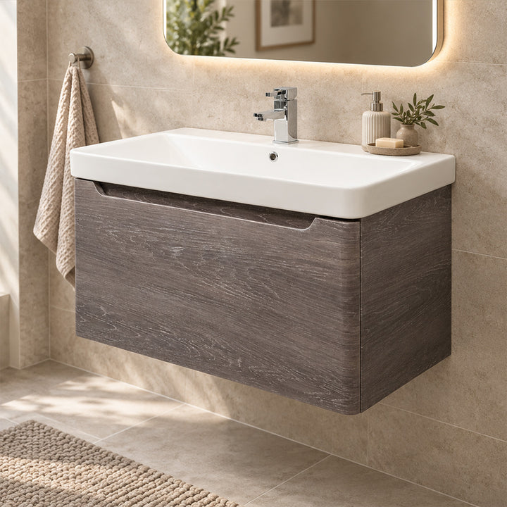 A modern wall-mounted bathroom vanity with a white rectangular sink and a dark wood-grain cabinet, shown installed against a beige tiled wall with a mirror, towel, and small decor items.