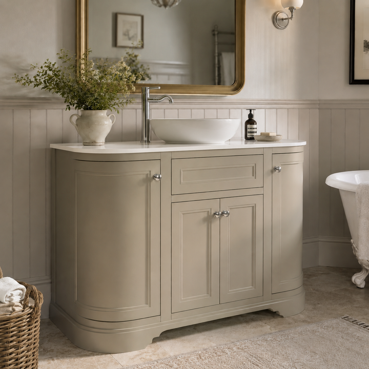 A Traditional floor standing bathroom vanity cabinet with a curved white countertop. The cabinet has a warm sand finish with silver knobs.