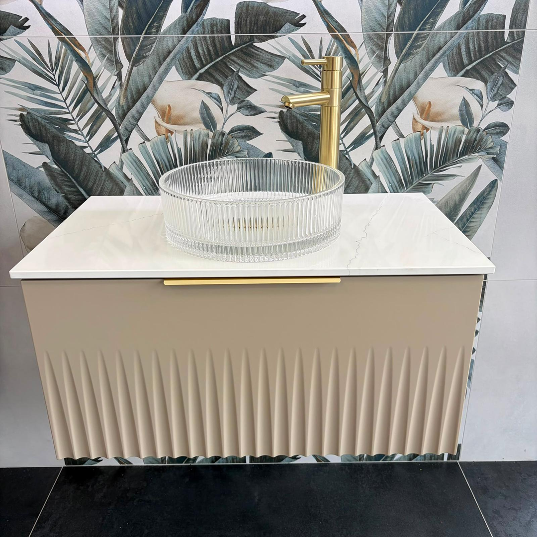 A modern bathroom vanity setup with a ribbed beige cabinet, white countertop, clear ribbed glass sink, and a gold tap